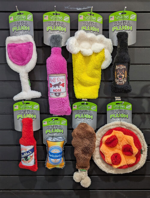 Wall display of dog toys shaped like bottles and a round pizza plush, including pink, yellow, and black plush bottles and a pizza rug on a black slat wall with green-packaged tags labeled 'Dura Plush'.