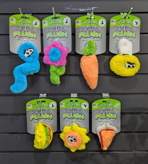 Colorful pet toys shaped like bones and animals hanging on a display with green and gray packaging labeled 'DURA PLUSH' in bright colors.