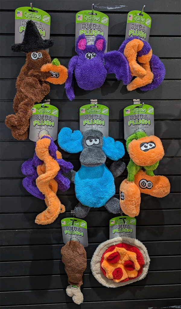 Colorful Cycle Dog Dura Plush pet toys hanging on a wall display in various shapes and colors (purple, blue, orange, brown) with packaging tags above each toy.