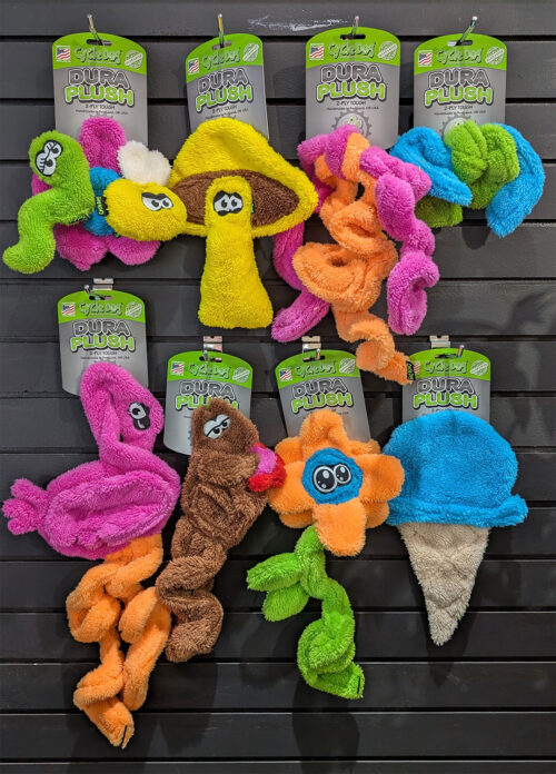 Colorful assortment of Cycle Dog Dura Plush plush dog toys hanging on a wall display, in bright green, pink, yellow, orange, blue, brown, and beige with product tags visible.
