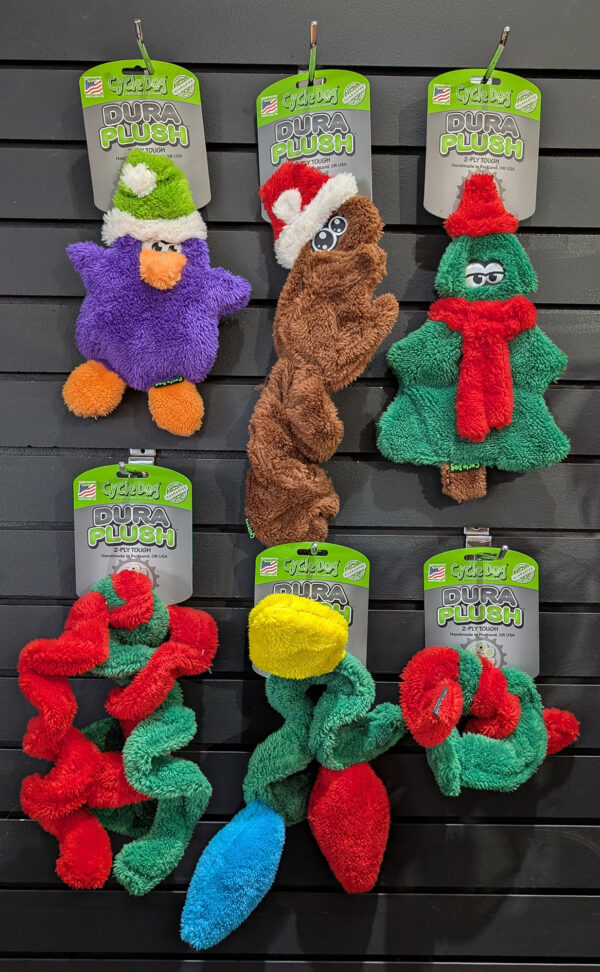Three Cycle Dog Dura Plush stuffed toys wearing festive hats hung on a display, with colorful garlands at the bottom.